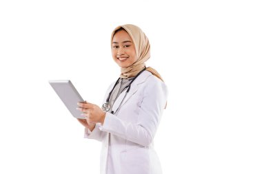 a beautiful doctor in doctor coat with hijab standing with smile while bring the tablet at her hand on the white background