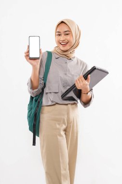 a beautiful student with hijab standing with the green bag at her shoulder while showing the phone at her right hand and bring the laptop at her left hand