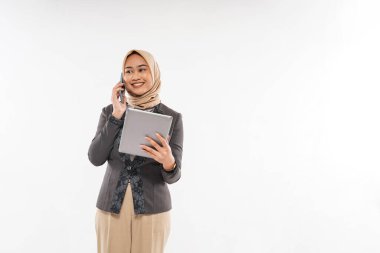 a young woman with hijab and gray blazzer standing with smile while calling someone on phone and bring the tablet at her hand on the white background