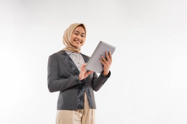 a beautiful woman with hijab and gray blazzer standing with smile and bring the tablet at her hand on the white background