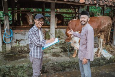 a moslem man smiling and pointing on the cow that standing outside the stable with the male farmer standing in front of him with the blue clipboard at his hand