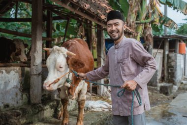 a moslem man with skullcap standing and holding the cows bridle that standing next to him