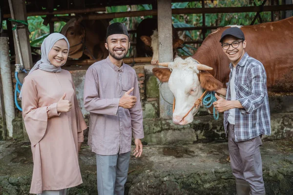 three adult standing with the cow in front of the cows stable with thumbs up and smiling