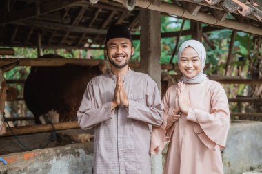 a man with skullcap and woman in hijab standing together with hands put together in front of the cows stable and smiling