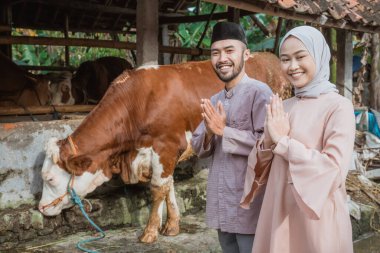 a moslem man and the woman with hijab standing with hands put together in front of the cows stable with the brown cow standing next to them