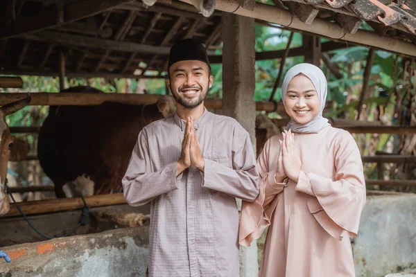 a man with skullcap and woman in hijab standing together with hands put together in front of the cows stable and smiling