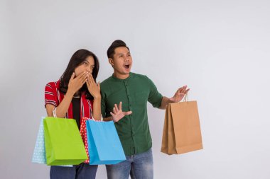 shocked asian couple looking away while bring the shopping bags at their hands on isolated background