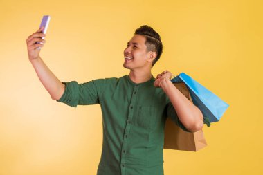asian man taking selfie photo while holding the shopping bags on his shoulder on orange isolated background