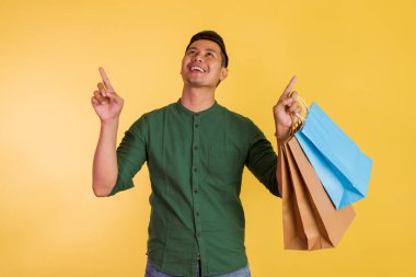 asian man looking up and pointing with his finger while holding the shopping bags on orange isolated background
