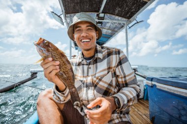 smiling Asian angler holding a grouper on a small fishing boat
