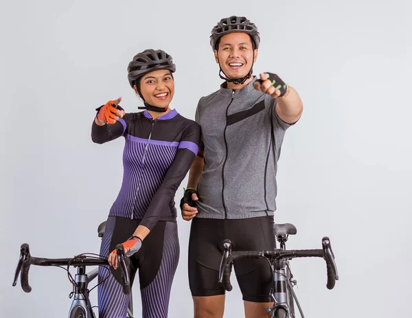 asian couple in cycling outfit standing with pointed finger beside their bicycles on isolated background