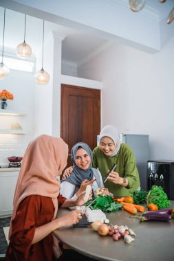 Three veiled women use cell phones while cooking together in the kitchen