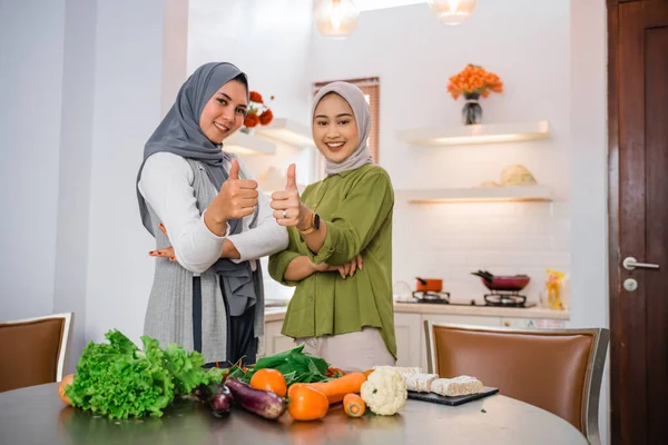 two women in hijabs smile with thumbs up while cooking in the kitchen