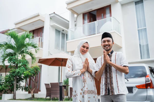 Asian Muslim husband and wife smiling with hand gestures wishing happy Eid in front of the house