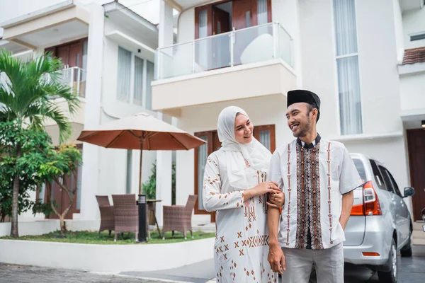 Asian Muslim husband and wife smiling intimately with hands holding in front of the house