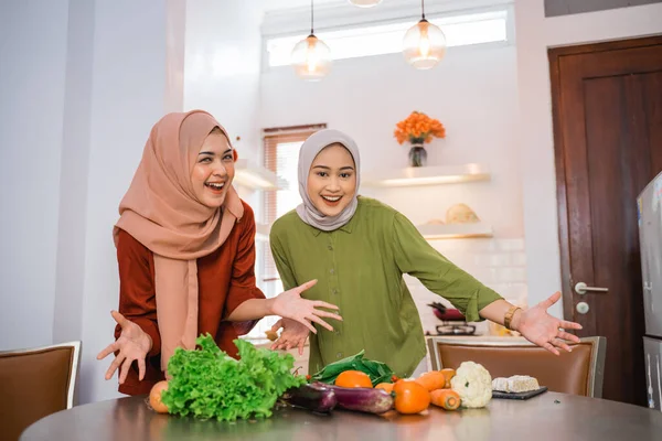 two veiled women smile with their hands presenting vegetables at the kitchen table
