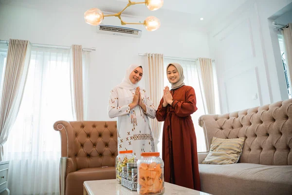 two women in hijabs smile with a gesture of wishing a happy Eid inside a house