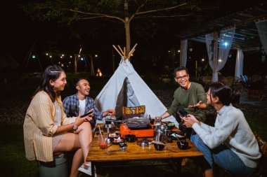 handsome guy in green long sleeved t shirt smiling and pointing on the woman in white knit sweater while hangout together at the camp site