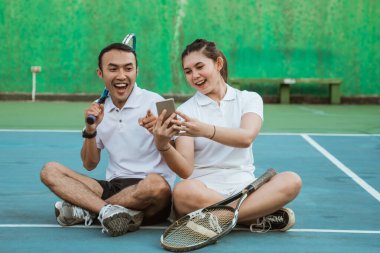 mixed doubles athlete together using mobile phone together while sitting on the tennis court
