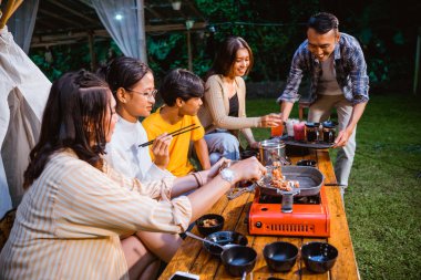 woman in orange stripped shirt taking the grilled beef from the grilled pan while the guy in plaid shirt put the drinks on the table