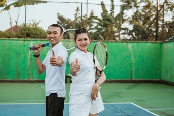 Smiling tennis athletes with thumbs up standing back to back on tennis court