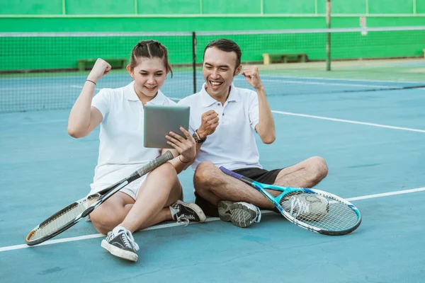 excited doubles tennis players sitting looking at pad screen together on tennis court