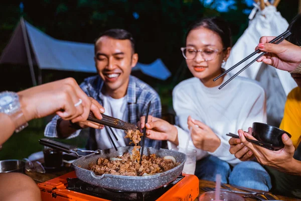 the guy in white t shirt and plaid shirt smiling showing his teeth while taking the grilled beef from the grilled pan using the chopsticks