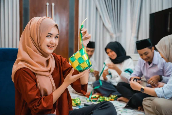 smiling Asian veiled woman holding woven ketupat ornaments for Eid decoration while gathering with friends