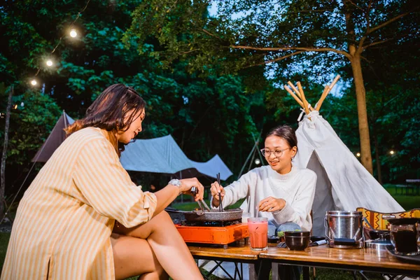 woman in white long sleeved t shirt with eye glasses helping the woman in orange stripped shirt cooking the beef on grill pan using the chopsticks