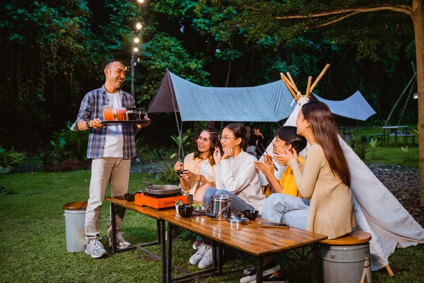 the guy in white t shirt and plaid shirt standing and bring the drinks using the tray for his friends that hangout together with him at the camp site