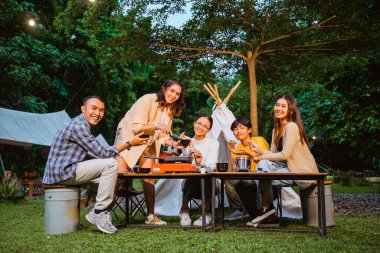 a group of people grilling beef using the grill pan happily at the camp site with the trees as the background