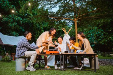 woman in orange shirt put the beef on the grill pan using the food tongs with her friends sitting around