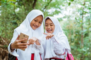 Üniformalı iki erkek ilkokul öğrencisi, yol kenarında yan yana durup izliyorlar ve telefonu gösterip gülüyorlar.