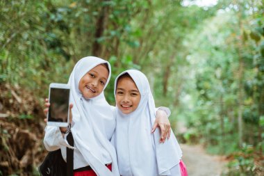 İki ilkokul öğrencisi, yol kenarında yan yana durmuş birbirlerine sarılıyorlar ve kameraya telefonu gösteriyorlar.