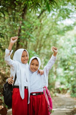 Üniformalı iki güzel ilkokul öğrencisi köy yolunda yan yana durur ellerini havaya kaldırır ve birbirlerine sarılırlar.