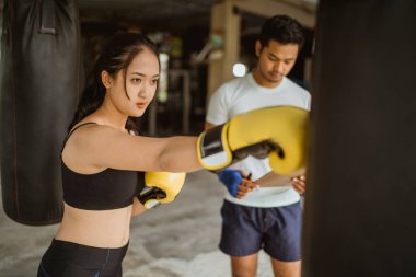 Erkek boks koçunun yanında duran bir bayan boksör boks stüdyosunda kum torbasını yumrukluyor.