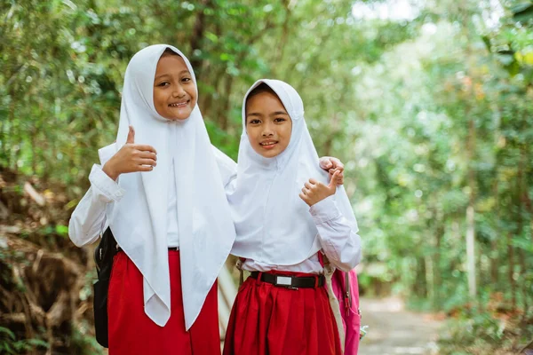 two beautiful elementary students in uniform standing together at the side of country road with thumbs up and hugging each other