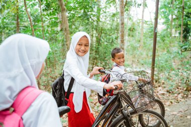 Üniformalı üç ilkokul öğrencisi birlikte kır yolunda yürüyorlar. Bisikletlerini getiriyorlar.