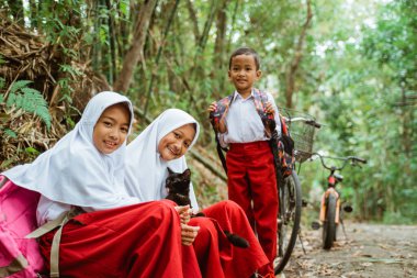 Üç ilkokul öğrencisi, kır yolunun kenarında siyah bir kediyle birlikte oturuyorlar.
