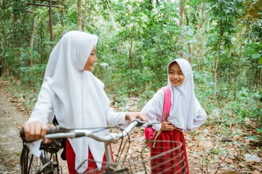 İki kız ilkokul öğrencisi birlikte yürüyorlar ve birbirlerine gülümsüyorlar. Biri bisikletini getiriyor, diğeri de elini beline koyuyor.