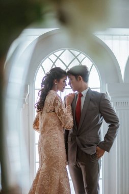 Asyalı gelin, arka plan olarak pencereyle yüz yüze dururken elini damadın omzuna koydu.