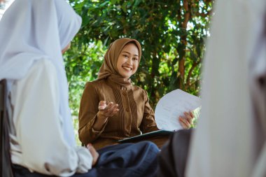 Açık hava sınıfında öğrencilere ders verirken jestli Asyalı öğretmen başörtüsü takıyor.
