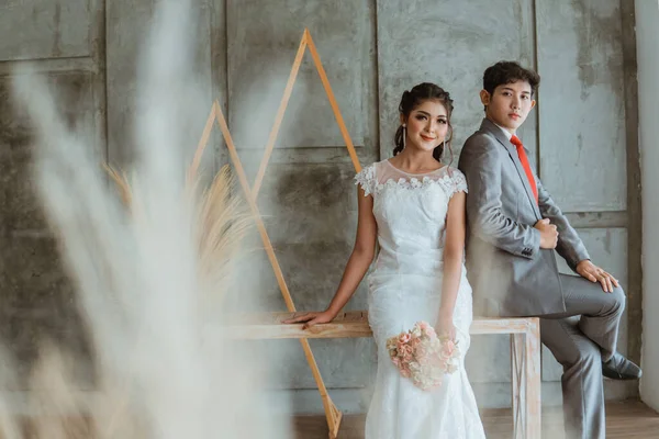 美丽的亚洲新娘 身穿白袍 手持花束 坐在新郎身旁 坐在摄影棚旁 — 图库照片