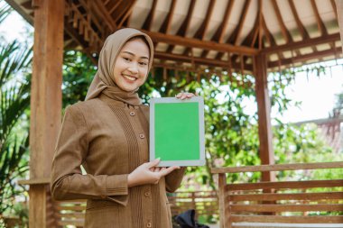 Tesettürlü Asyalı bayan öğretmen taraça arkaplanlı bir tablet ekranı gösteriyor