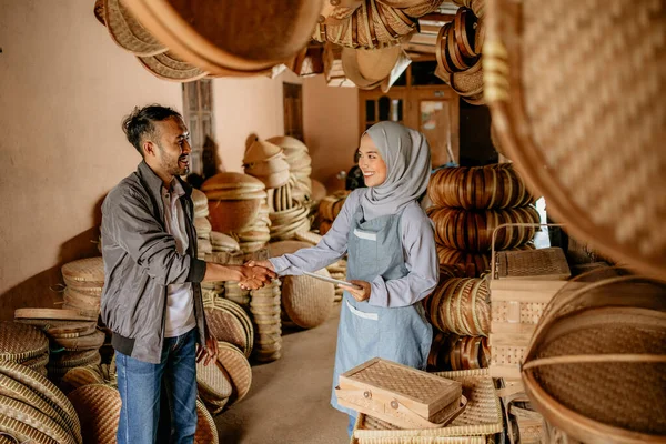 Güzel, genç Müslüman kadın, dokunmuş bambu ürünleri atölyesinde müşteriyle tokalaşıyor.