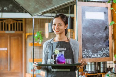 Önlüklü bayan barista kahvede tepsiyle içkiyi getirirken gülümsüyor.