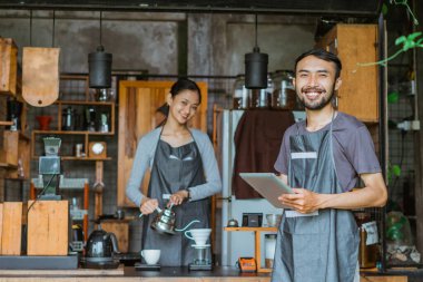 Önlüklü erkek barista arka planda bayan baristayla dijital tableti tutuyor.