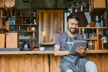 Barın önünde otururken dijital tablet kullanan önlüklü bir erkek barista.