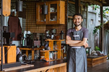 Önlüklü yakışıklı barista elleri çapraz duruyor ve bar masasının yanında gülümsüyor.