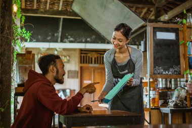 Kahve dükkanındaki kadın baristanın gösterdiği menü defterini gösteren erkek müşteri.